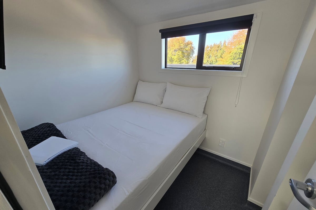 A small, single-bed room with a window, a door, and a carpeted floor.