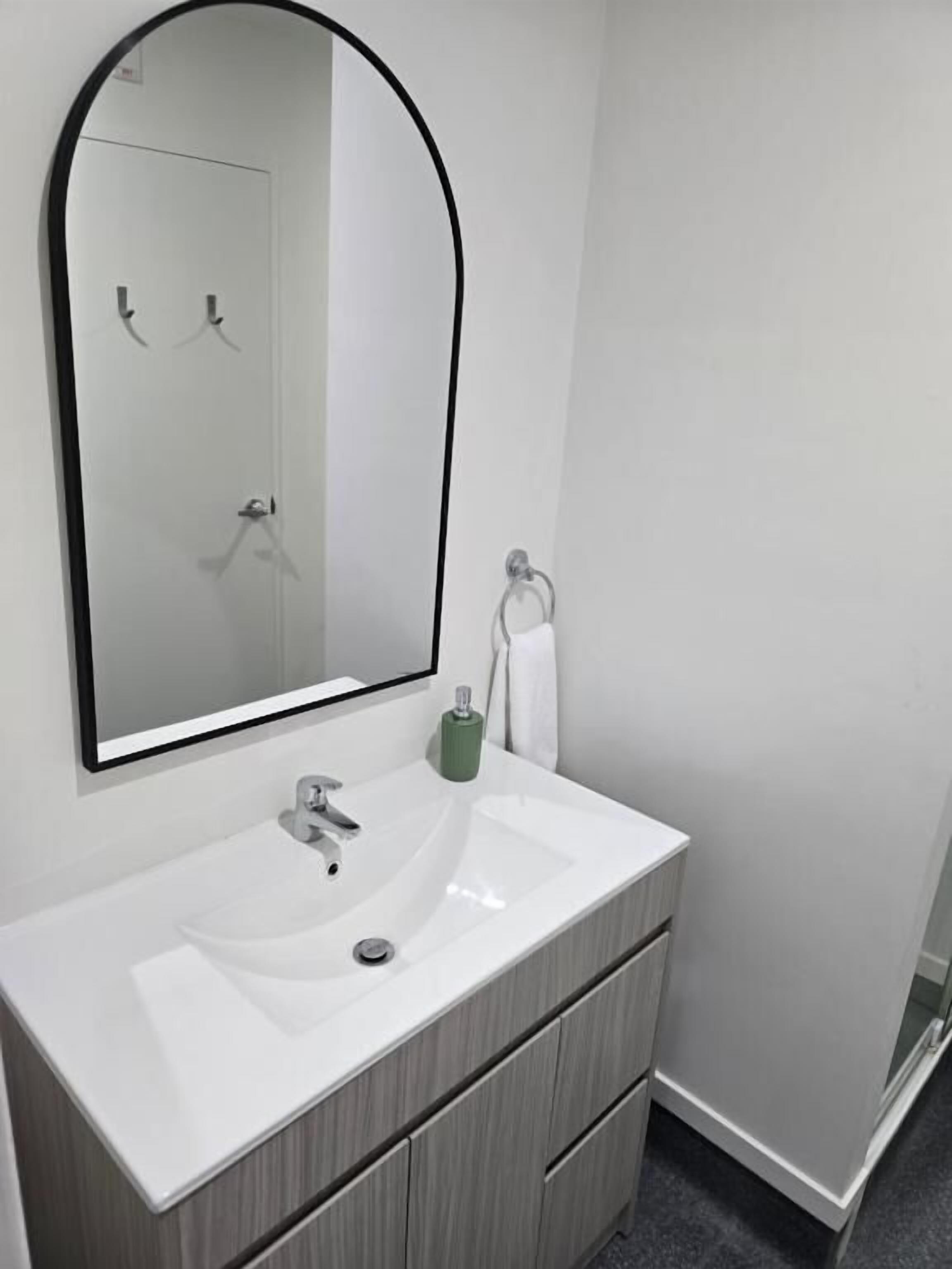 Un bagno con un lavandino bianco, uno specchio con cornice nera, un dispenser per sapone verde e un porta asciugamani.