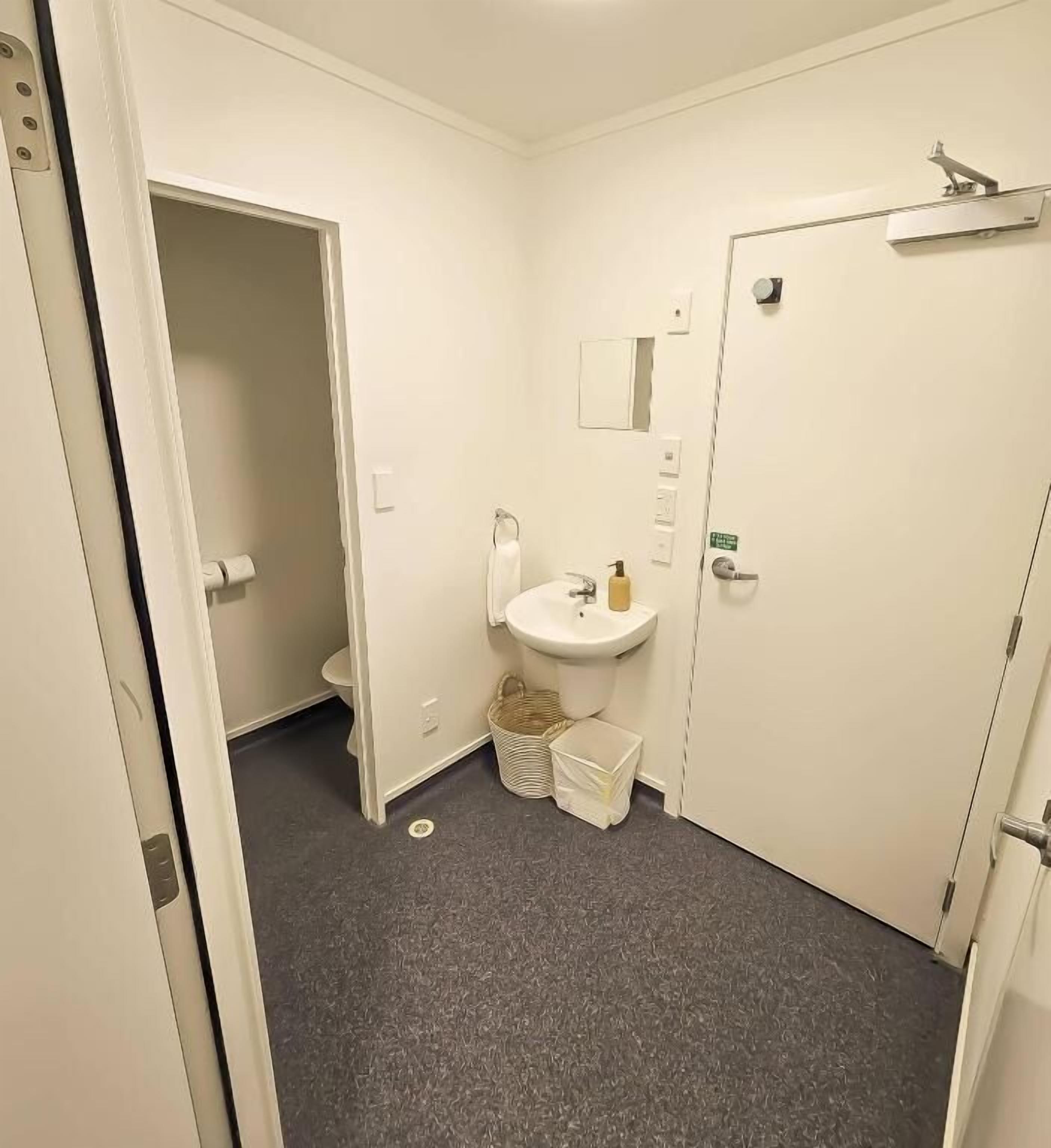 Un baño pequeño y funcional con inodoro, lavabo y espejo sobre él. Se observa una puerta a la derecha y un cubo de basura al lado del lavabo.