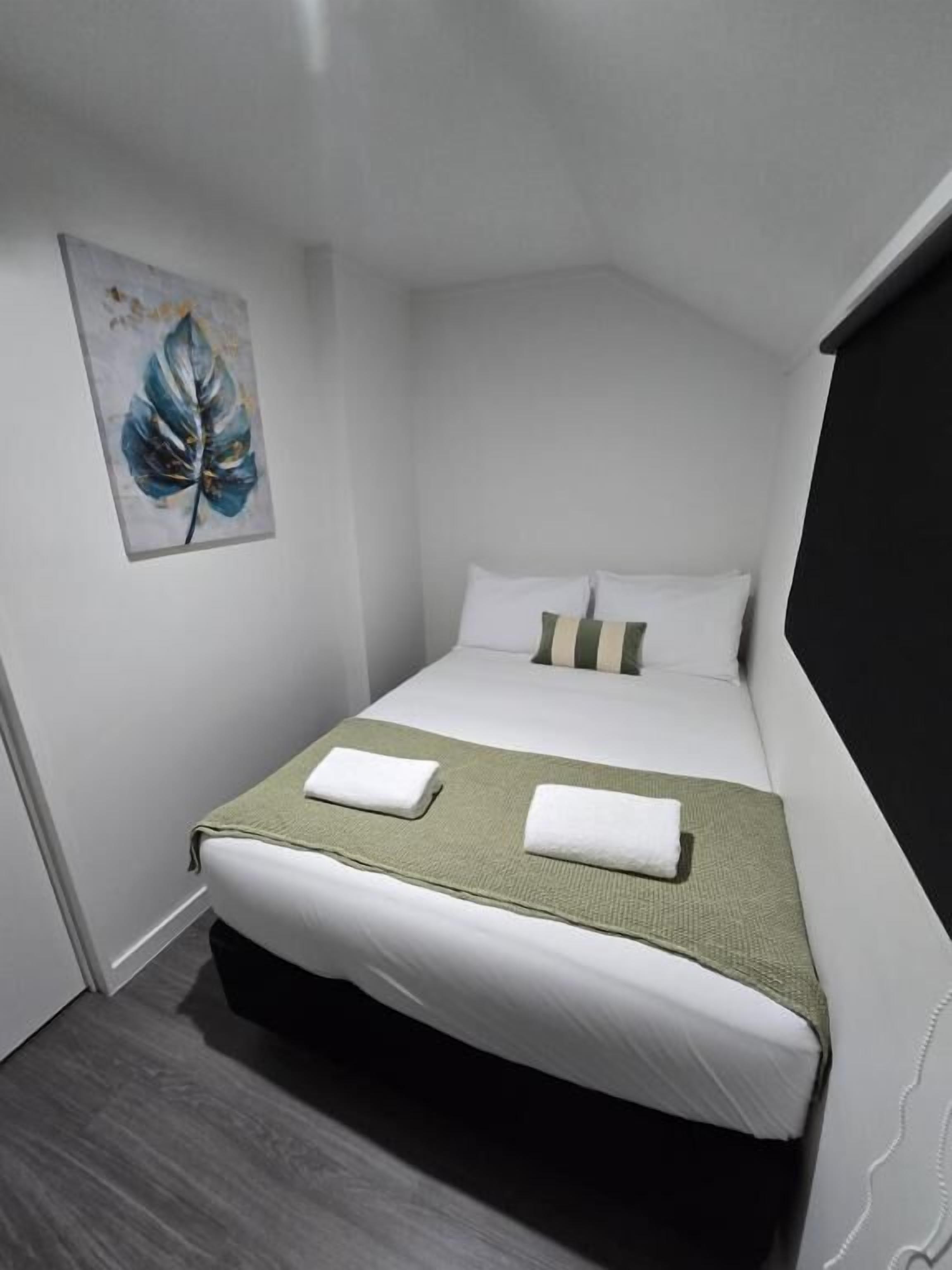 A neatly made bed with white linens and a green throw blanket, a pillow with a striped pillowcase, and a single artwork of a leaf on the wall.