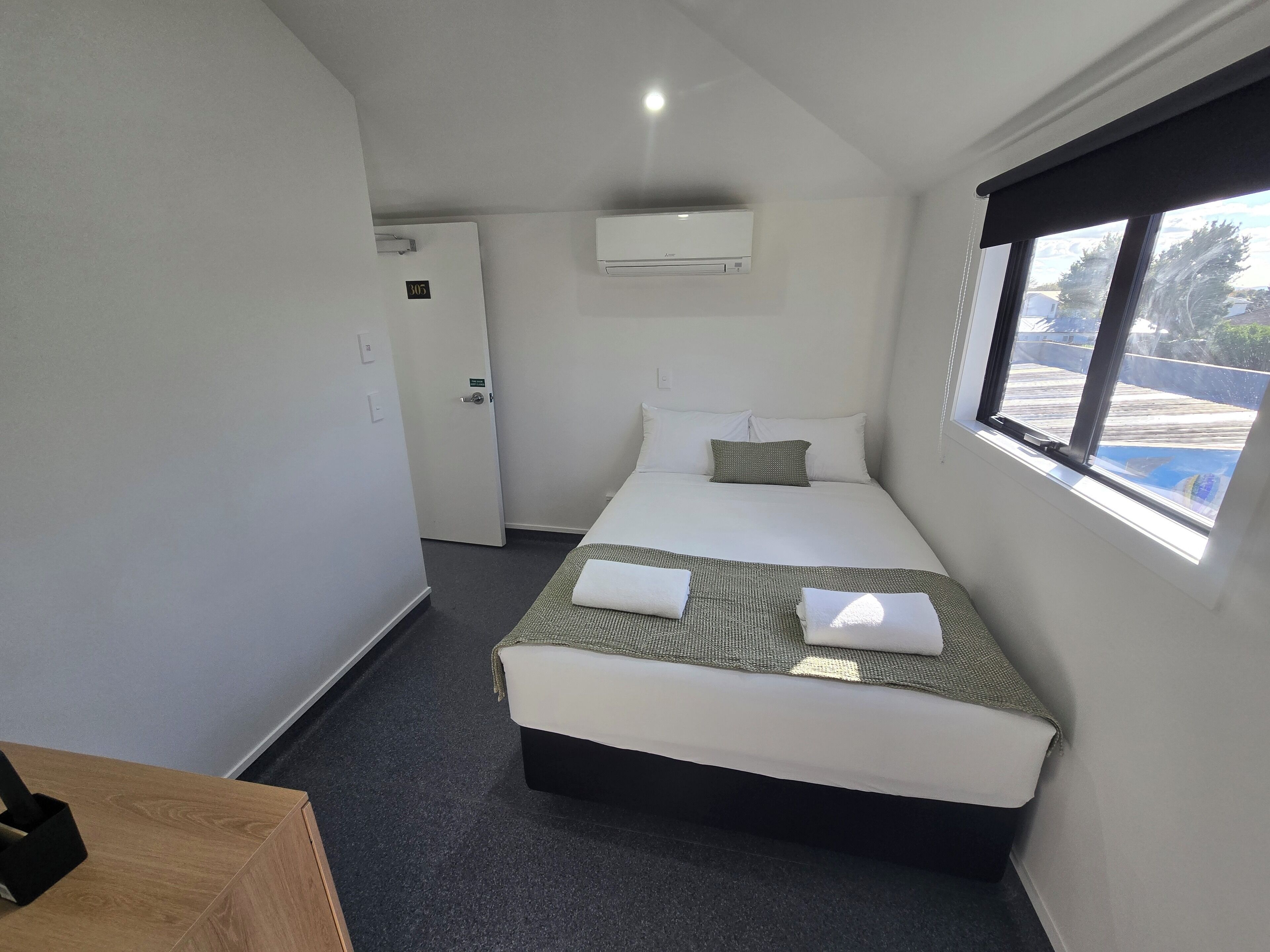 Une chambre d’hôtel avec un lit simple, une petite table, un appareil de climatisation, une fenêtre donnant sur un paysage agréable et une porte portant un numéro.