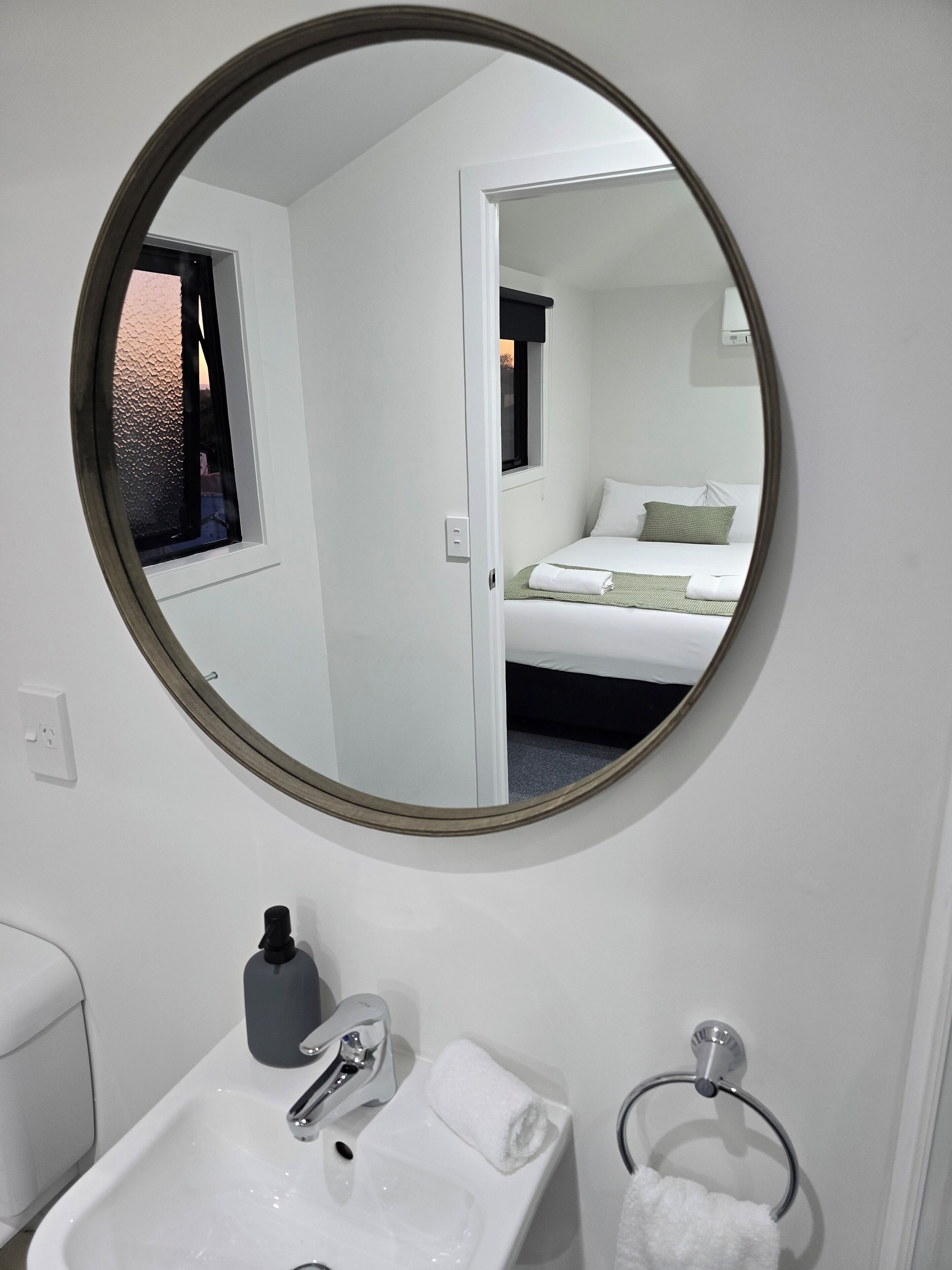 Un bagno con uno specchio rotondo, un lavandino, un porta asciugamani e una finestra che dà su una camera da letto.