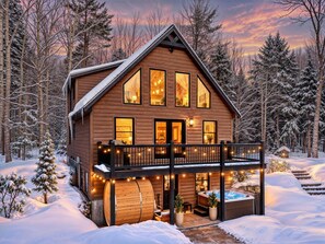 A wooden cabin with a balcony, a hot tub, and a deck surrounded by snow.