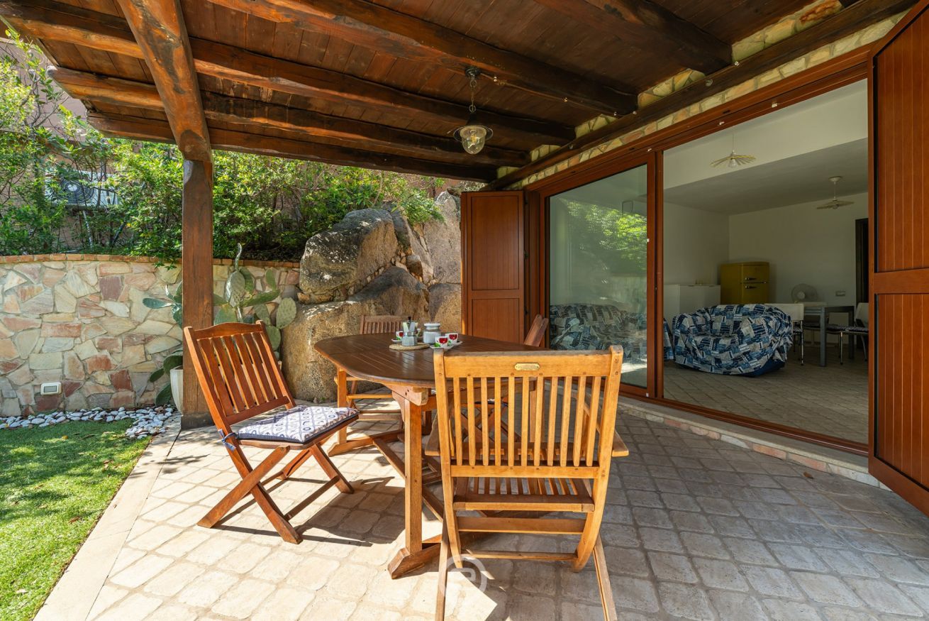 Une terrasse extérieure avec une table et des chaises en bois, un mur en pierre et une grande porte-fenêtre coulissante donnant sur un espace intérieur.
