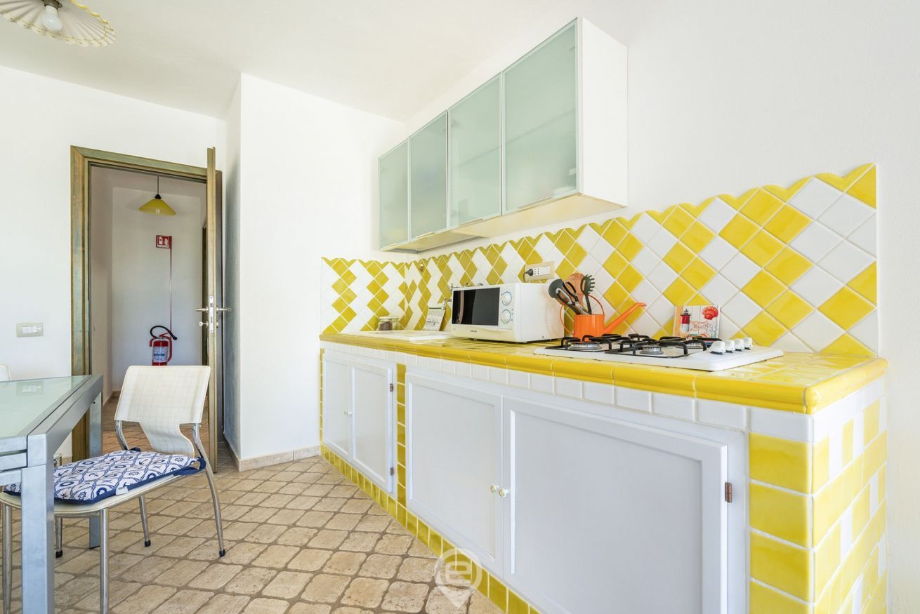 A kitchen with a yellow and white tiled backsplash, a microwave, a gas stove, and a chair.