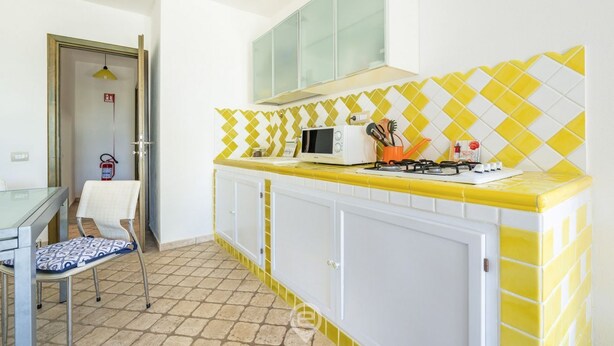 A kitchen with a yellow and white tiled backsplash, a microwave, a gas stove, and a chair. - Alessandra’s Garden Hideaway (Simius)