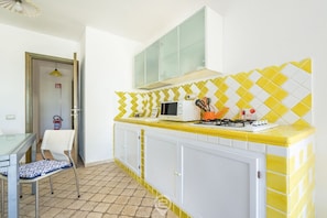 Une cuisine avec un plan de travail recouvert de carreaux jaunes et blancs, un four à micro-ondes, une cuisinière à gaz et une chaise.