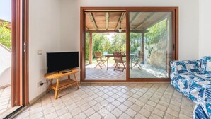 A room with a TV on a wooden stand, a patterned sofa, and a sliding glass door leading to an outdoor patio with a table and chairs. - Alessandra’s Garden Hideaway (Simius)