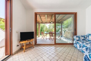 A room with a TV on a wooden stand, a patterned sofa, and a sliding glass door leading to an outdoor patio with a table and chairs. - Alessandra’s Garden Hideaway (Simius)