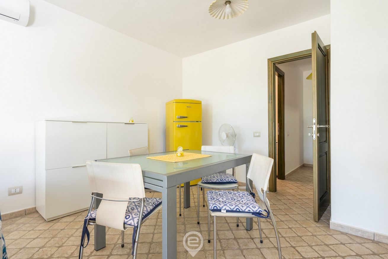 Une pièce de taille modeste, avec une table à plateau de verre, des chaises blanches, un appareil électroménager jaune et une porte donnant sur une autre pièce.