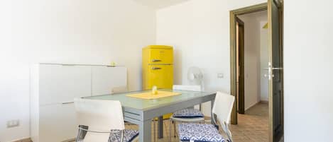 A compact room with a glass-top table, white chairs, a yellow appliance, and a door leading to another room.