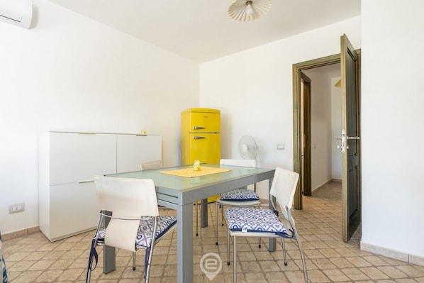 Une pièce de taille modeste, avec une table à plateau de verre, des chaises blanches, un appareil électroménager jaune et une porte donnant sur une autre pièce.
