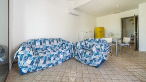 A room with two covered sofas, a small table with chairs, a yellow water dispenser, and a white air conditioning unit. - Alessandra’s Garden Hideaway (Simius)