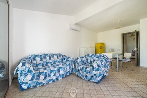A room with two covered sofas, a small table with chairs, a yellow water dispenser, and a white air conditioning unit. - Alessandra’s Garden Hideaway (Simius)