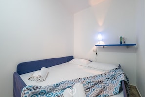 Un lit simple avec une tête de lit bleue, une couverture à motifs et une serviette enroulée posée sur le lit. Il y a une étagère fixée au mur, sur laquelle se trouvent une lampe et un livre.