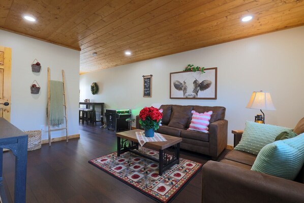 Un salon chaleureux avec un plafond en bois, un canapé marron, une table basse ornée d’un vase de fleurs, un tapis à motifs et un tableau encadré représentant une vache accroché au mur.