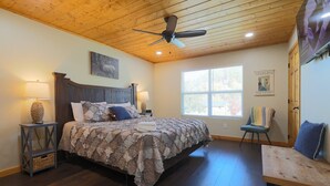 Een slaapkamer met een houten plafond, een bed met een bedovertrek met patroon, een nachtkastje met een lamp, een stoel en een raam met uitzicht op bomen.