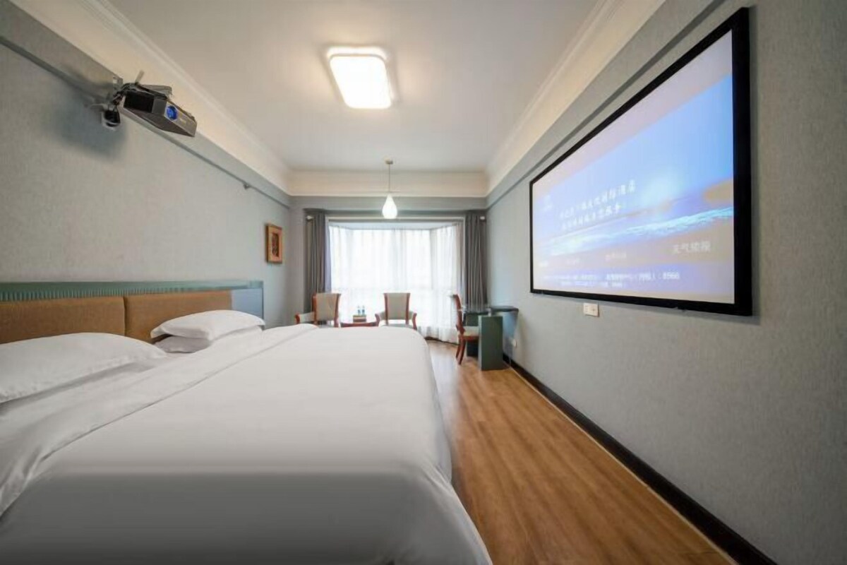 Une chambre d’hôtel avec un grand lit, un écran de projection mural et une vue sur l’extérieur par la fenêtre.