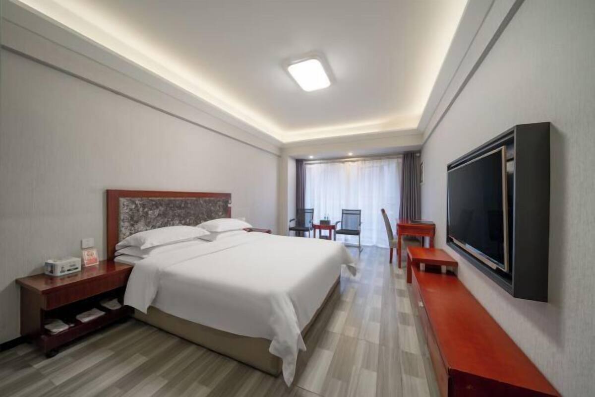 Ein Hotelzimmer mit einem großen Bett, einem an der Wand befestigten Fernseher, einem Schreibtisch mit Stuhl und einem kleinen Tisch mit Stühlen.
