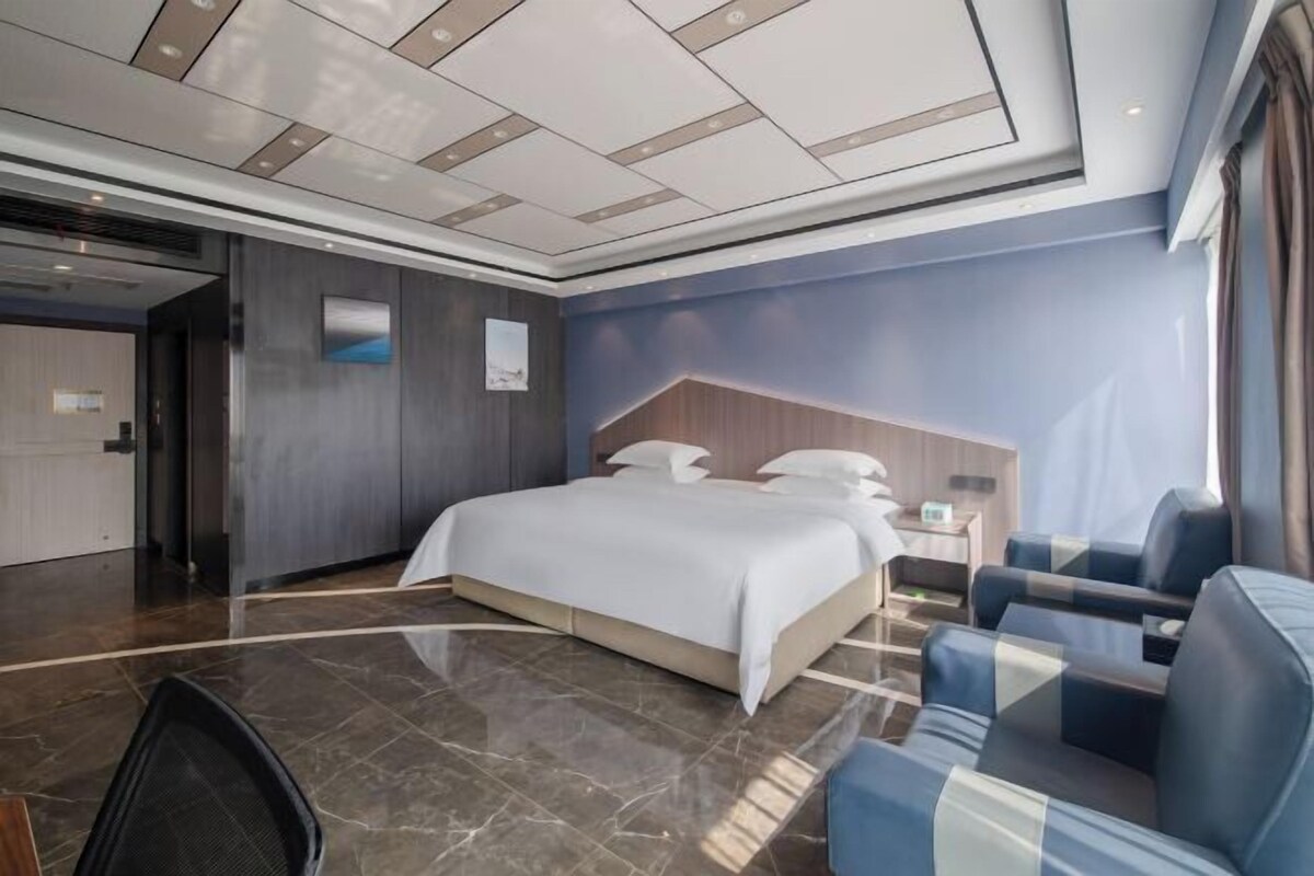 A modern hotel room with a large bed, a seating area with blue sofas, a wooden headboard, and a large window providing natural light.