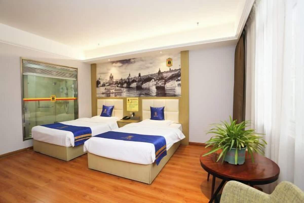 A hotel room with two beds, a glass partition, a cityscape mural, a small table with a plant, and a chair.