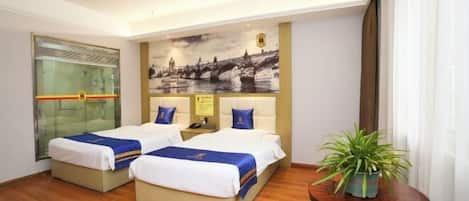 Quarto de hotel com duas camas, divisória de vidro, mural com vista da cidade, uma pequena mesa com planta e uma cadeira.
