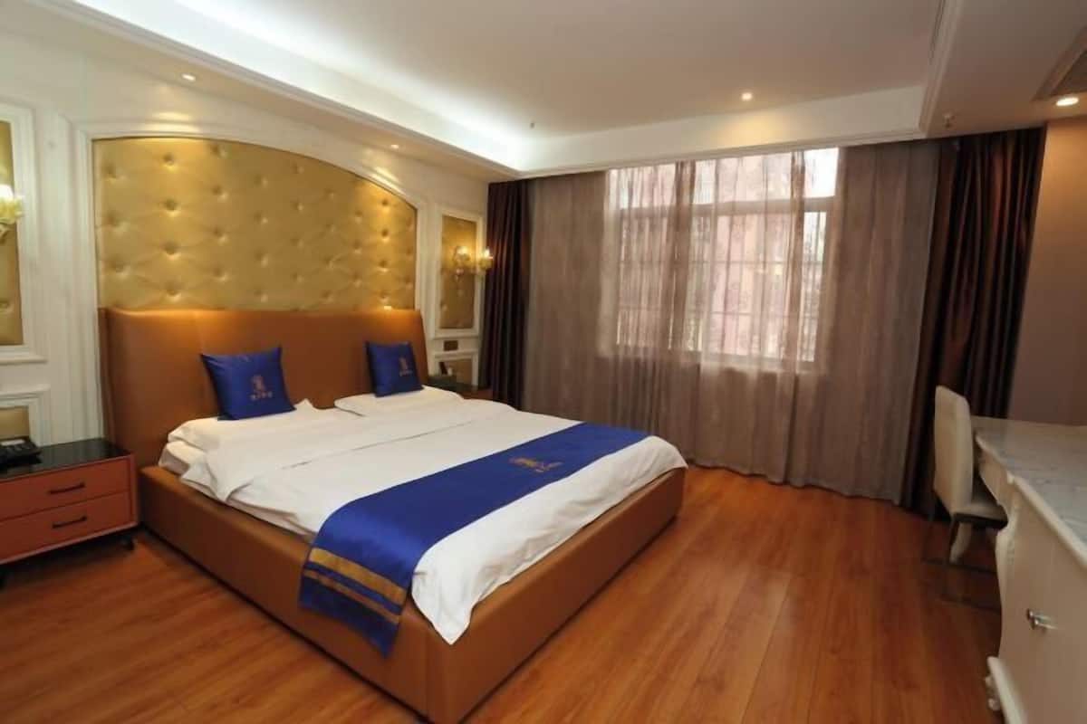 A hotel room with a large bed, a wooden floor, a headboard with a tufted design, and a window with curtains.