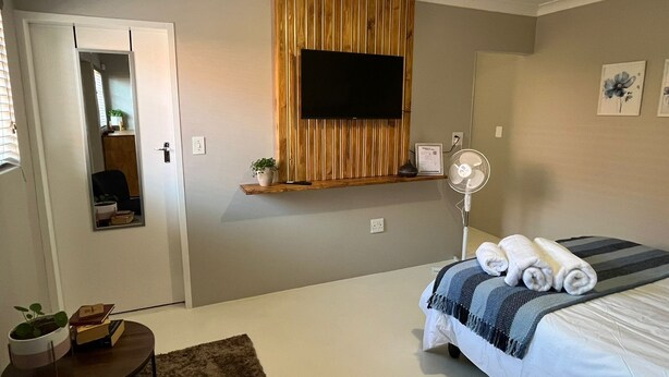 A bedroom with a bed, a TV mounted on a wooden panel, a bedside table with a plant, and a small table with books.