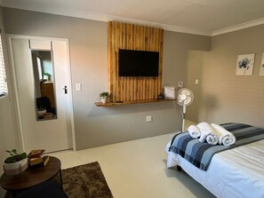A bedroom with a bed, a TV mounted on a wooden panel, a bedside table with a plant, and a small table with books.