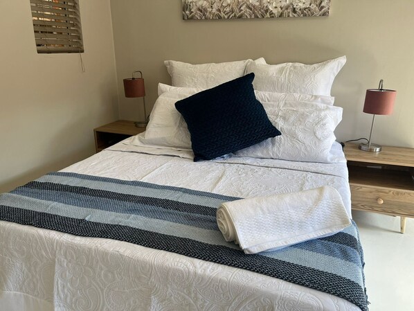 A neatly made bed with white linens and a dark blue pillow, flanked by two bedside tables with lamps, in a room with a simple, clean design.