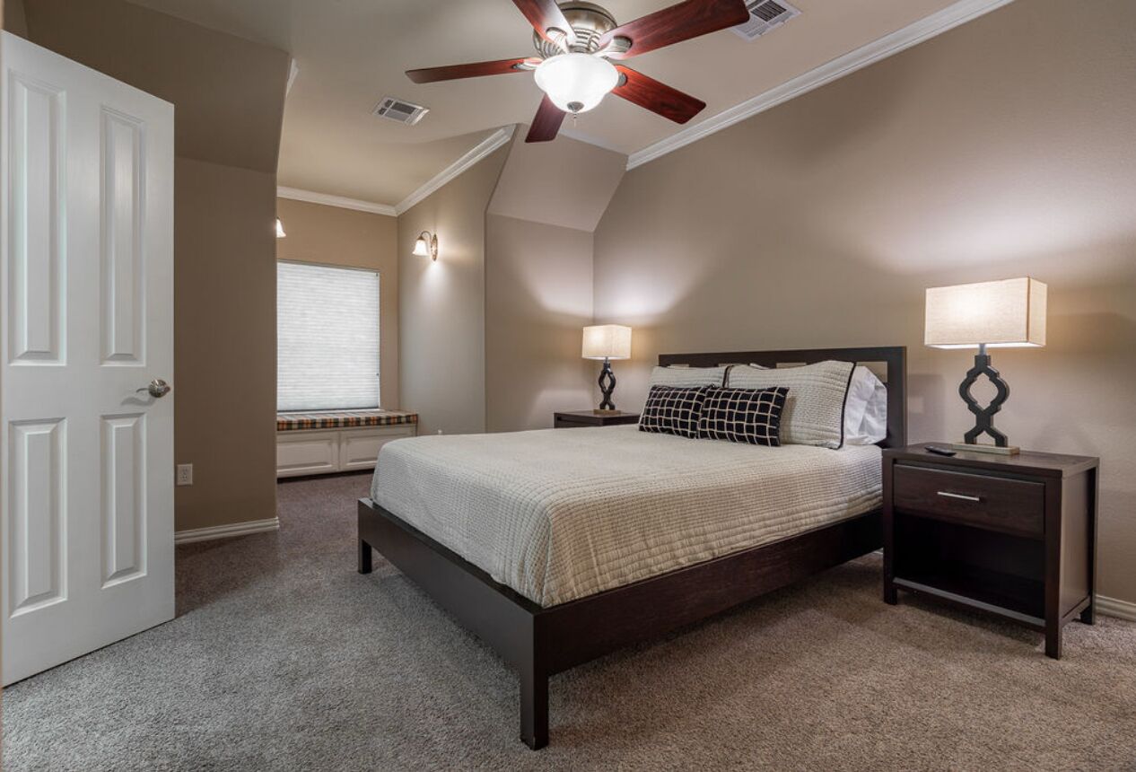A bedroom with a bed, two bedside tables with lamps, a ceiling fan, and a window with blinds.