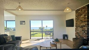 A spacious living room with a large sliding glass door leading to a balcony with a view of the sea. There is a dark-colored sofa, a small round table, and a TV mounted on the brick wall. - BEACH FRONT - Sunflower Beach House Pohara (Pōhara)