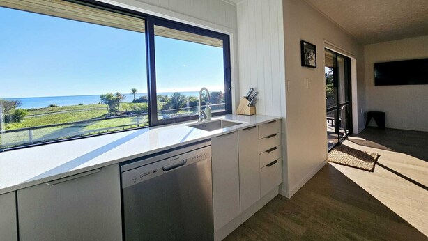 A modern kitchen with a large window overlooking a scenic view, a dishwasher, and a minimalist design. - BEACH FRONT - Sunflower Beach House Pohara (Pōhara)