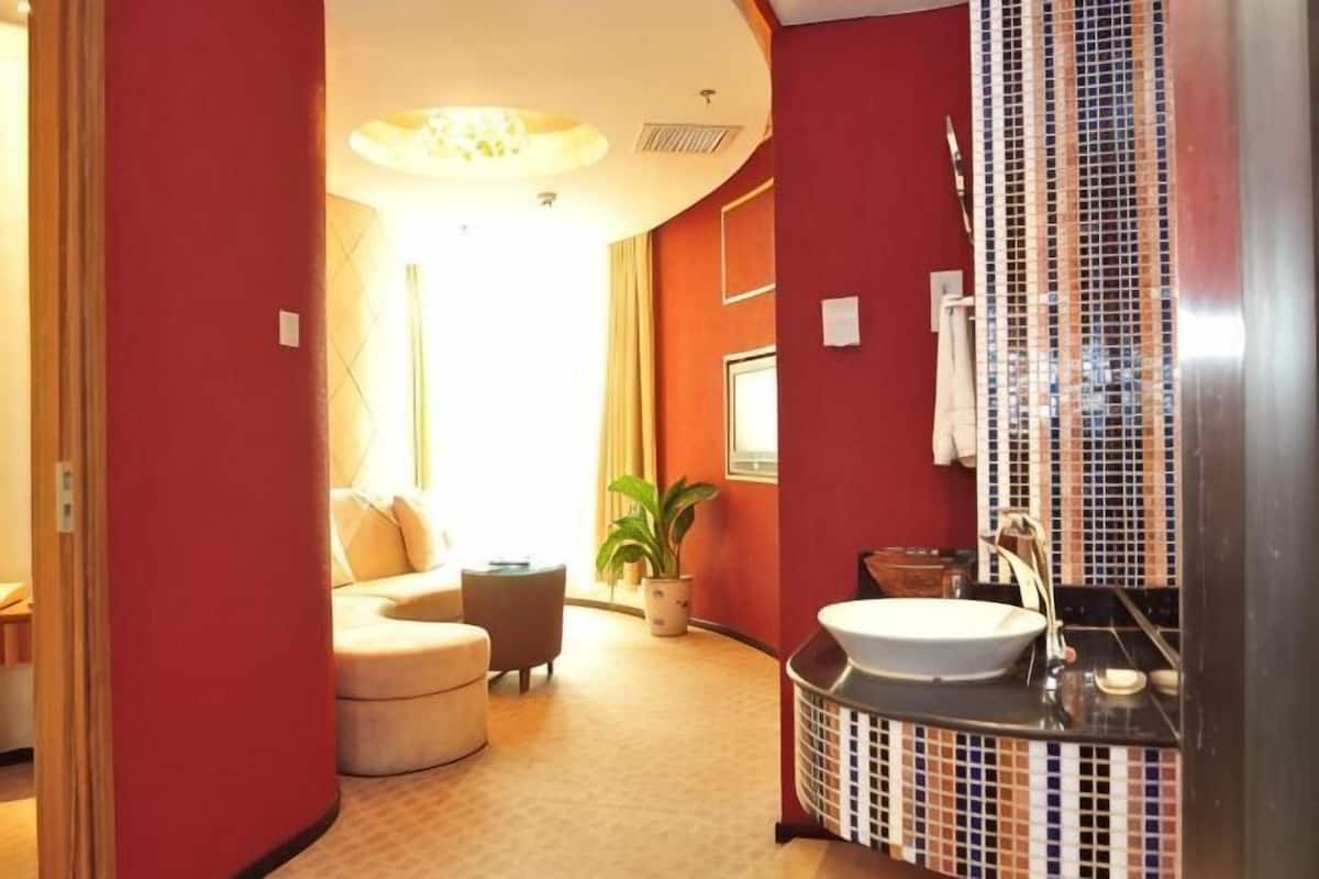 A hotel room with a curved sofa, a small table, a potted plant, a round sink, and a tiled shower area.