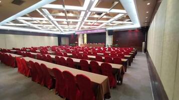 Sala de conferências com fileiras de mesas e cadeiras, palco com um púlpito e teto com padrão geométrico.