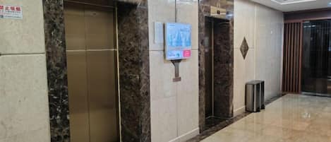 A hotel lobby with marble flooring, two elevators, a trash bin, and a digital information kiosk.