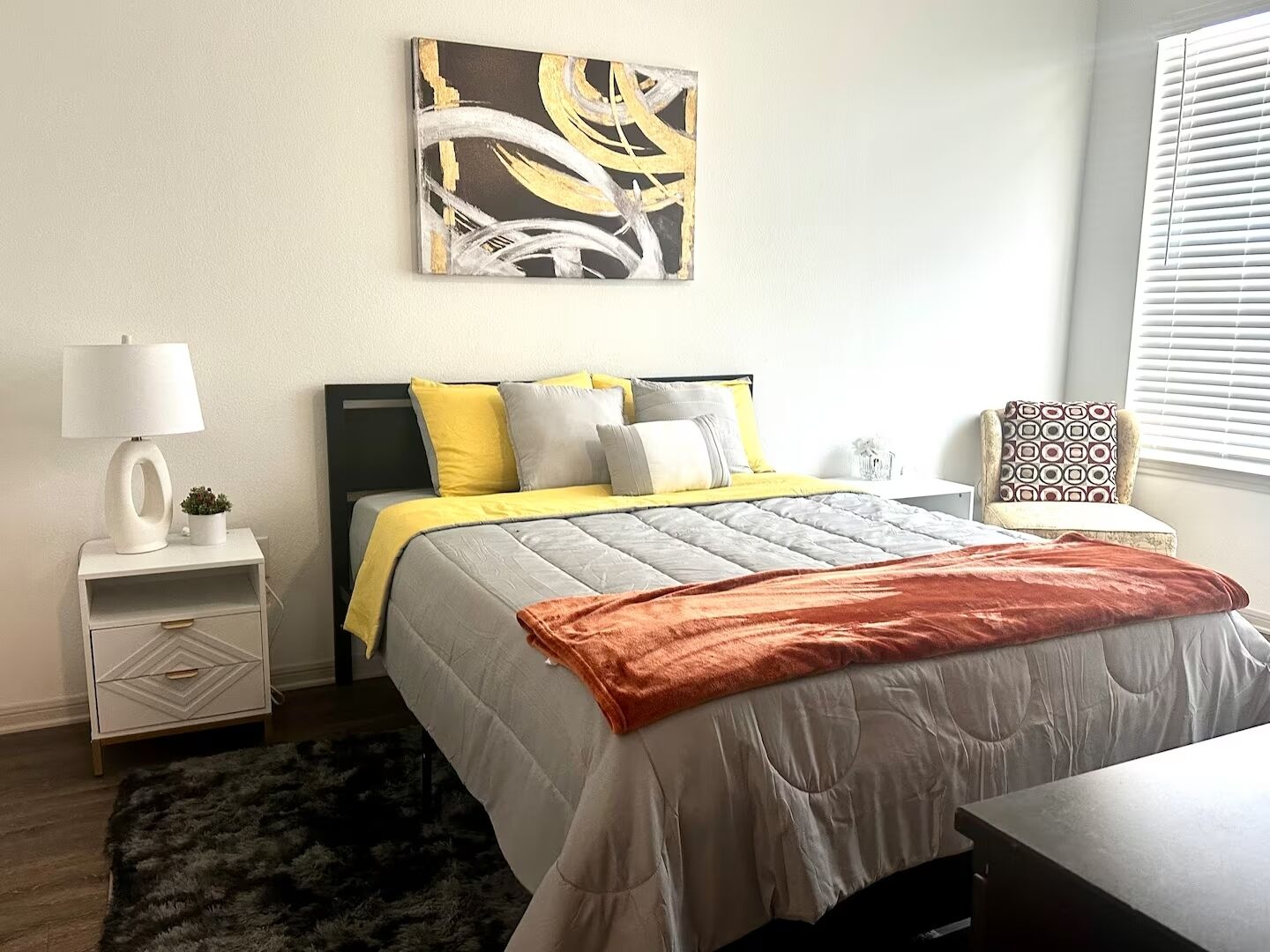 Un dormitorio moderno con cama, mesitas de noche, lámpara, una silla y una ventana con persianas.