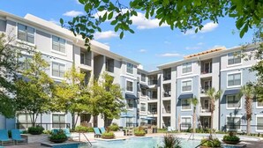 A modern apartment building with a central swimming pool, outdoor seating, and palm trees.