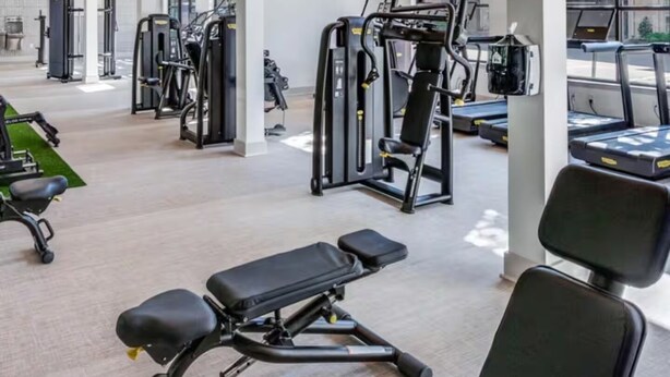 A spacious gym with various exercise equipment, large windows, and a high ceiling.