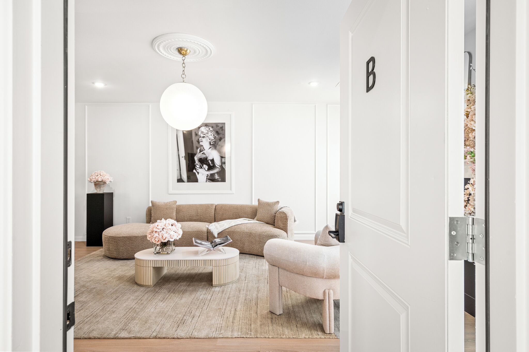 Un salon moderne avec un canapé beige, une table basse ronde et un fauteuil blanc. Un grand portrait en noir et blanc orne le mur, et une porte blanche porte la lettre « B ».