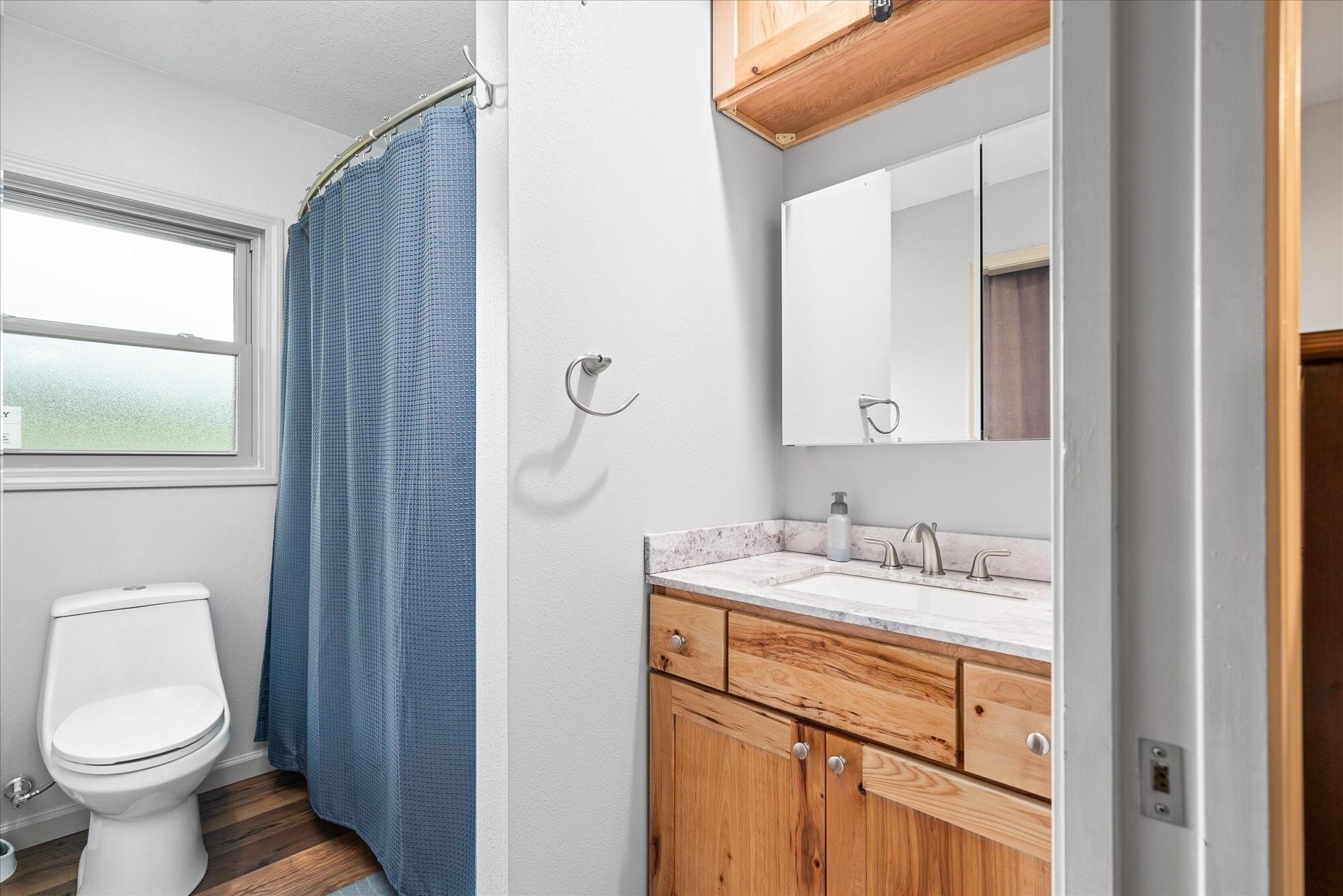 Une salle de bain avec des toilettes blanches, un rideau de douche bleu, un meuble-lavabo en bois avec un plan de travail en marbre et un miroir au-dessus du lavabo.