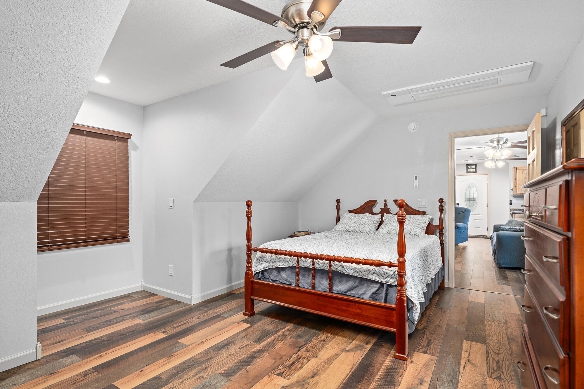 Une chambre avec un plafond en pente, un lit à baldaquin en bois, un ventilateur de plafond et une vue sur le salon attenant.