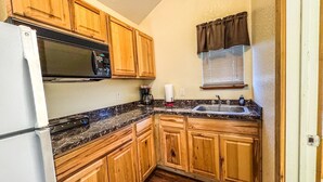A compact kitchen with wooden cabinets, a microwave, a refrigerator, a sink, and a window with blinds. - Cozy cabin with AC and WiFi in peaceful Sterling City.  Your small town adventure awaits! (Sterling City)
