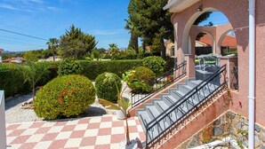Una escalera conduce a un espacio al aire libre cubierto con piso a cuadros, rodeado de setos cuidados y una palmera.