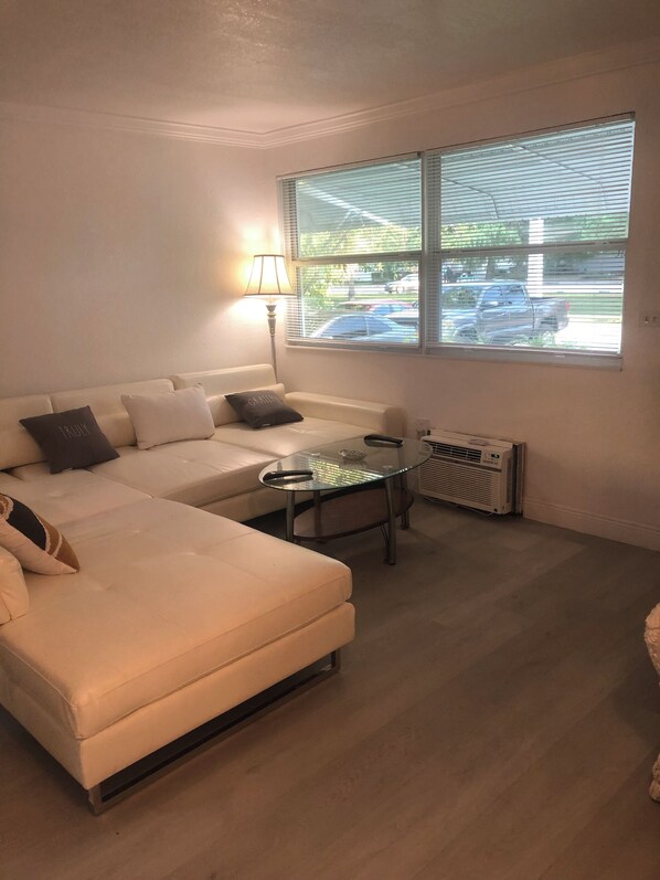 A cozy living room with a sectional sofa, a glass coffee table, and a window with blinds. - Charming 2 bedroom lake apartment in lovely Hallandale Beach with paddleboat!!! (Hallandale Beach)