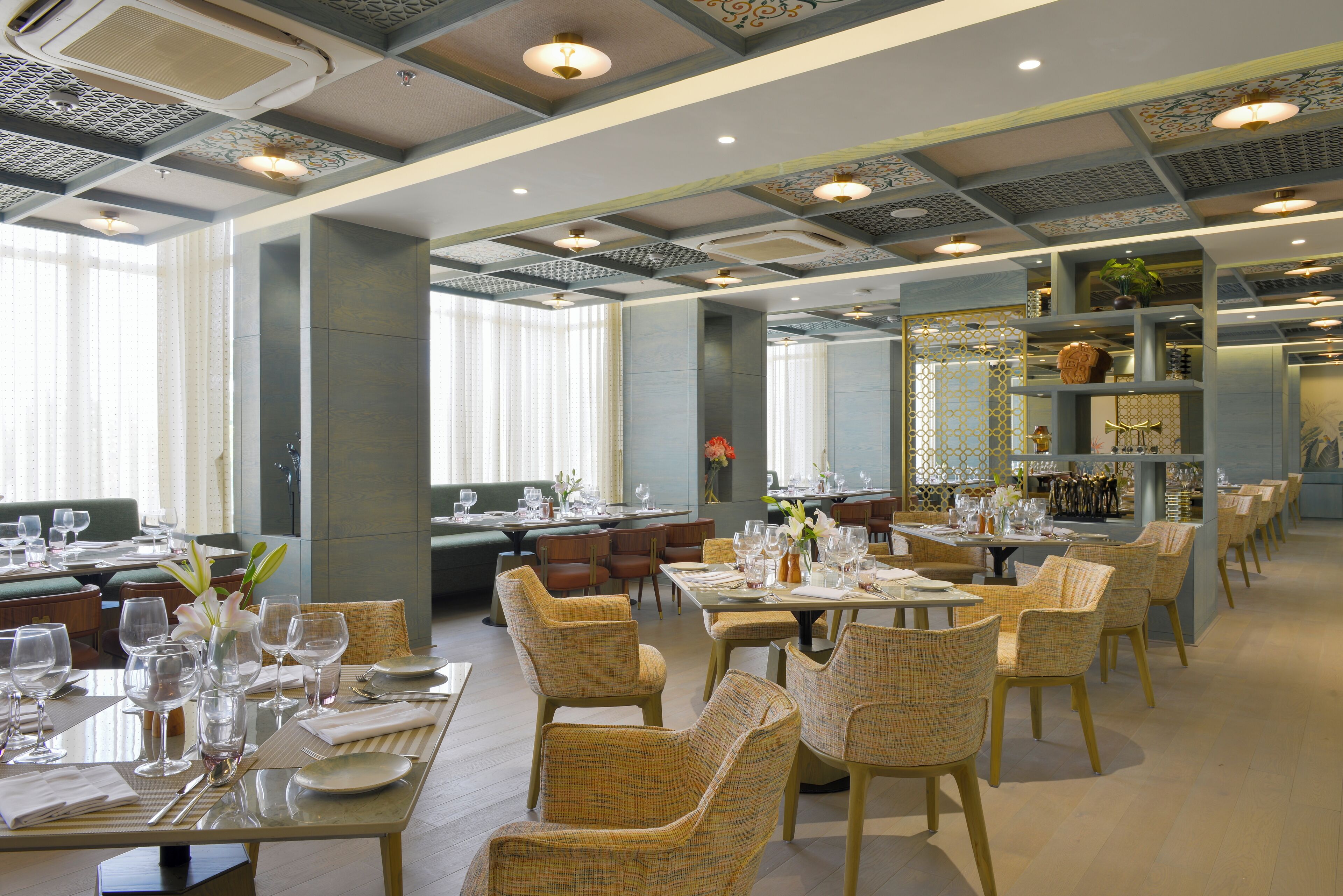 A spacious dining area with multiple tables and chairs, large windows with sheer curtains, and a decorative ceiling with intricate patterns.