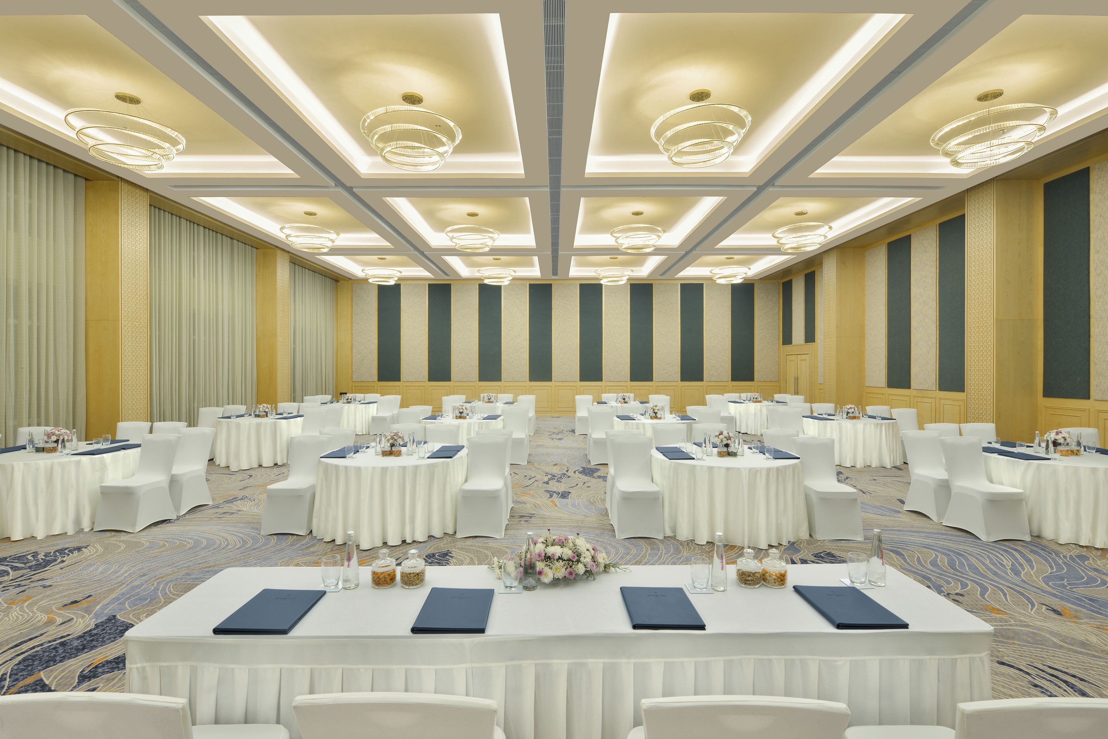 A large, well-lit conference room with round tables and white chairs, featuring a patterned carpet and a decorative ceiling.