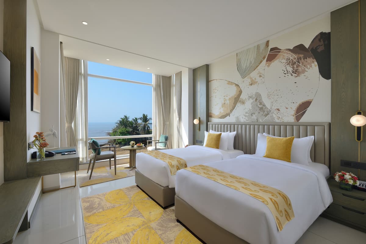 A hotel room with two beds, a desk, a chair, a TV, and a view of the ocean.