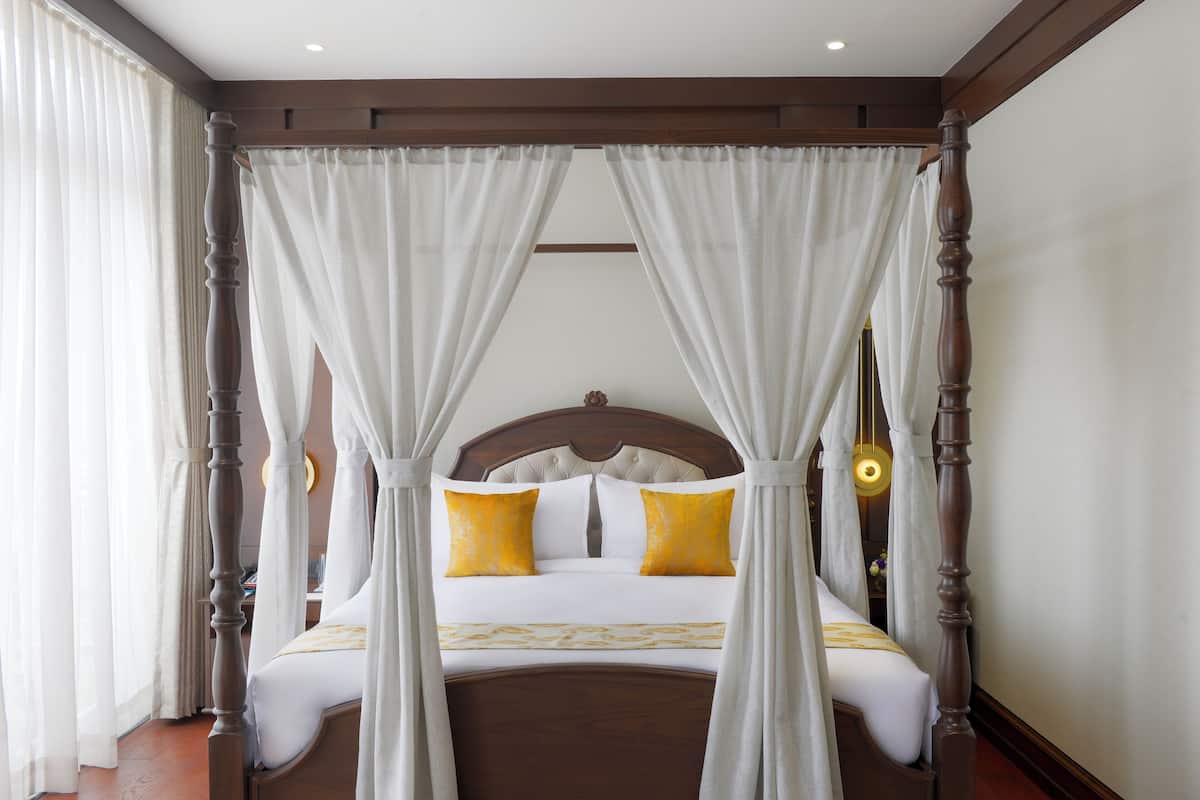 A four-poster canopy bed with white and yellow bedding, wooden headboard, and a nightstand with a lamp.