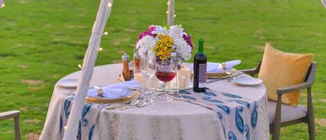 Mesa de comedor al aire libre con mantel blanco, una botella de vino y un centro de mesa floral.